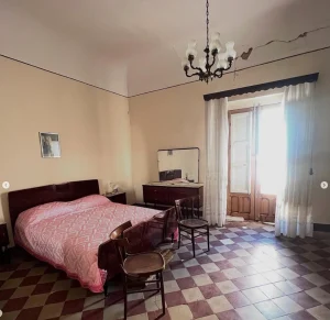Chambre principale d’un appartement en Sicile avec lit double, meubles anciens et balcon lumineux.